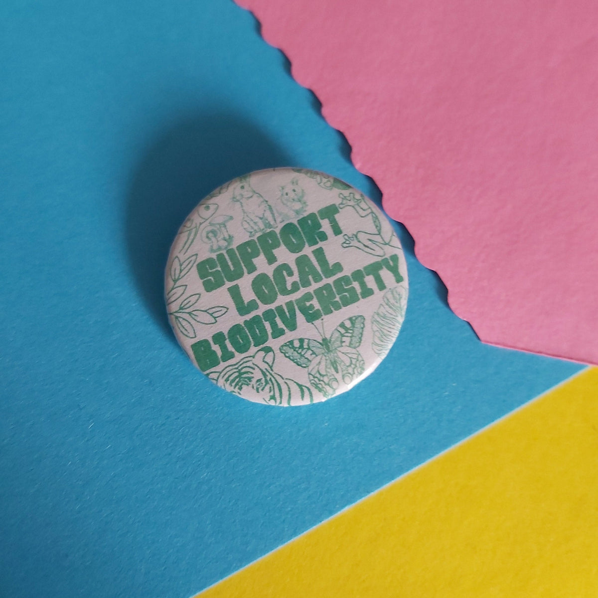 A round pinback button badge with the text 'SUPPORT LOCAL BIODIVERSITY' in green, surrounded by light green nature illustrations, placed on a colorful background of blue, pink, and yellow paper.