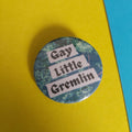 A round button pin with the text 'Gay Little Gremlin' in black gothic font on white banners, set against a dark green and blue swamp pattern. The pin is photographed on a background split diagonally between yellow and blue.