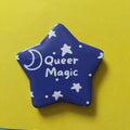 A glossy blue star-shaped pin badge with white stars, dots, a crescent moon, and the words 'Queer Magic' in white script, placed on a bright yellow background.