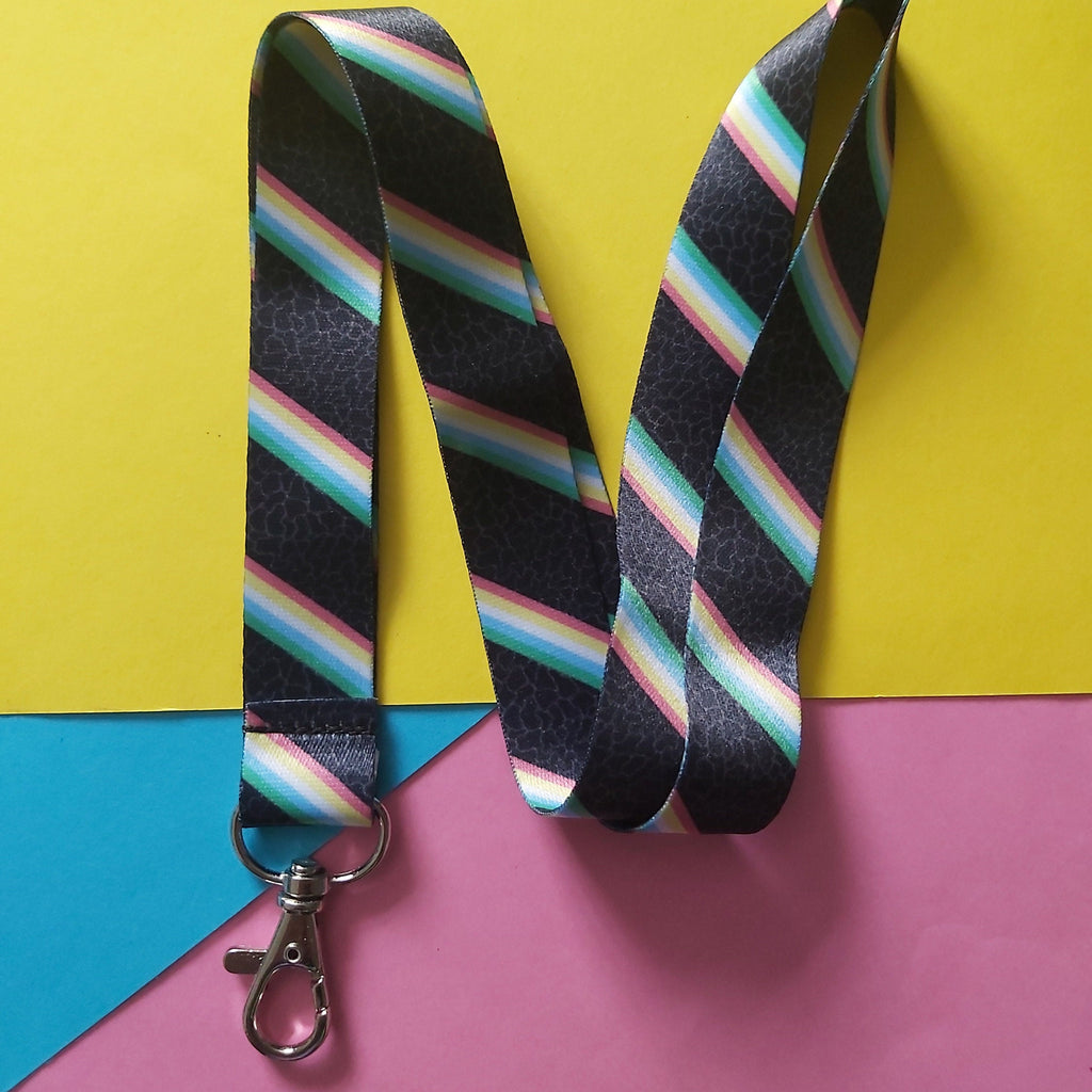 A black lanyard with diagonal rainbow stripes and a silver lobster clasp, laid flat on a colourful background of yellow, pink, and blue paper.