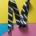 A black lanyard with diagonal rainbow stripes and a silver lobster clasp, laid flat on a colourful background of yellow, pink, and blue paper.