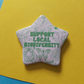 A star-shaped pin badge featuring green line art illustrations of animals, plants, and the text 'SUPPORT LOCAL BIODIVERSITY' in green on an off-white background, set against a bright yellow surface.