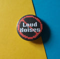 A round pin badge with a black background, red pixelated prohibition symbol, and white text reading 'Loud Noises', photographed on a split yellow and blue background.