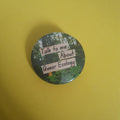 A round button pin featuring a forest scene with three beige text banners that read 'Talk to me about queer ecology