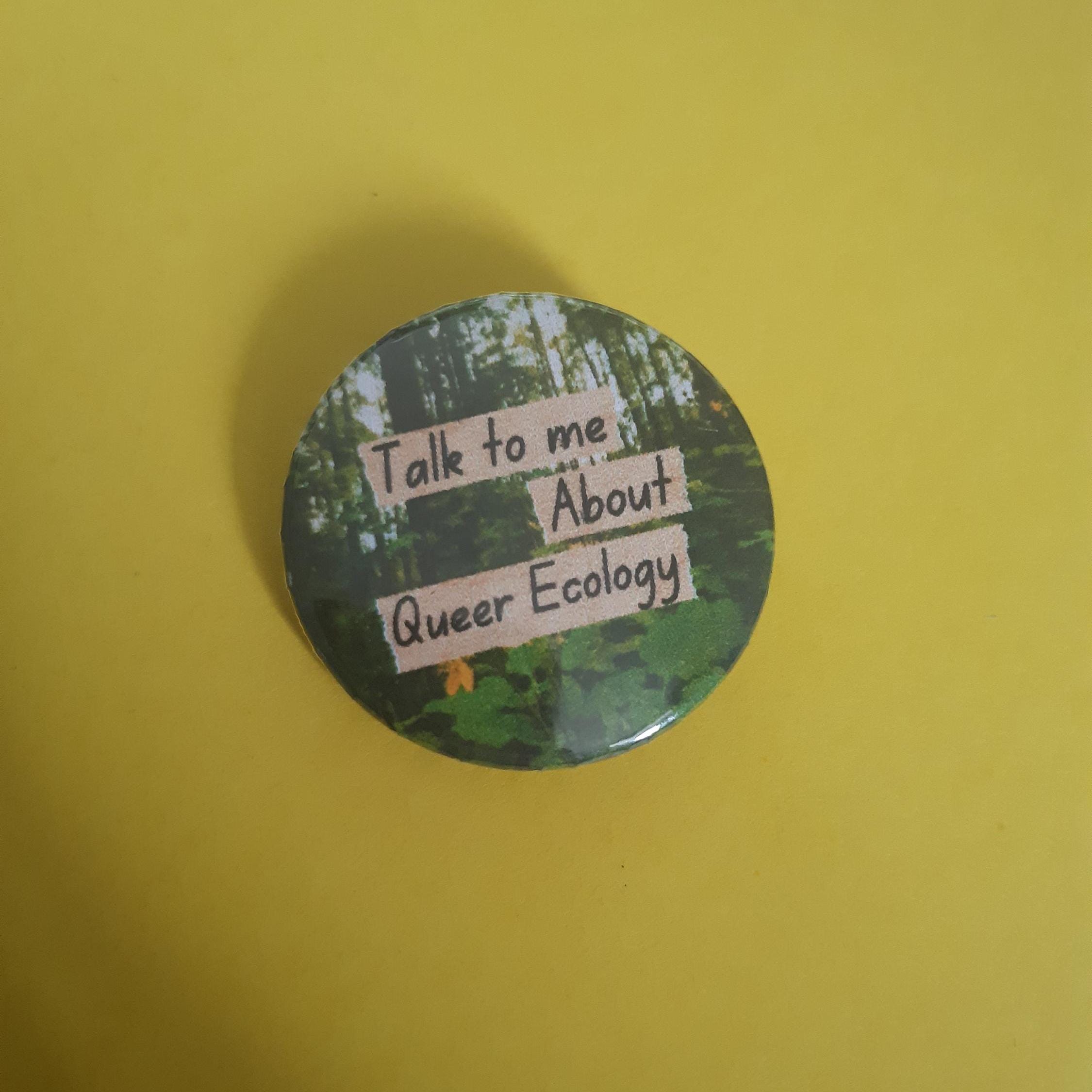 A round button pin featuring a forest scene with three beige text banners that read 'Talk to me about queer ecology