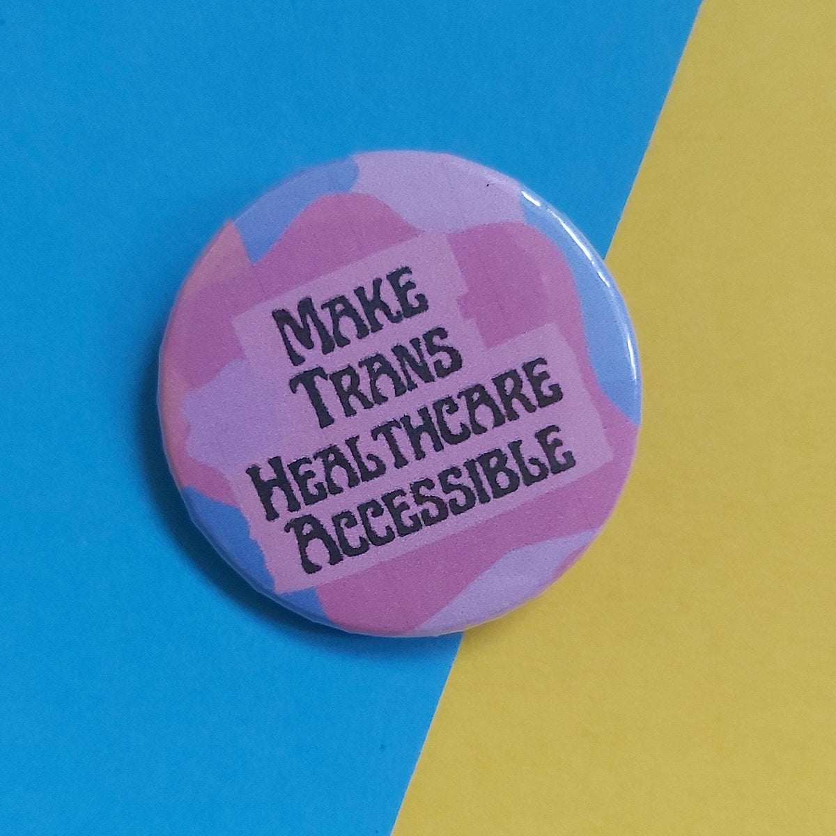 A round pinback button badge with a pink and blue abstract pattern, featuring the text 'MAKE TRANS HEALTHCARE ACCESSIBLE' in black. The button is photographed on a background split diagonally between bright blue and yellow.