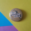 A round pinback button badge with a brown kraft-paper background and black western-themed graphic, featuring the text 'GAY COWBOY CLUB' on white strips. The badge is photographed on a colourful background split diagonally into yellow, blue, and pink sections.