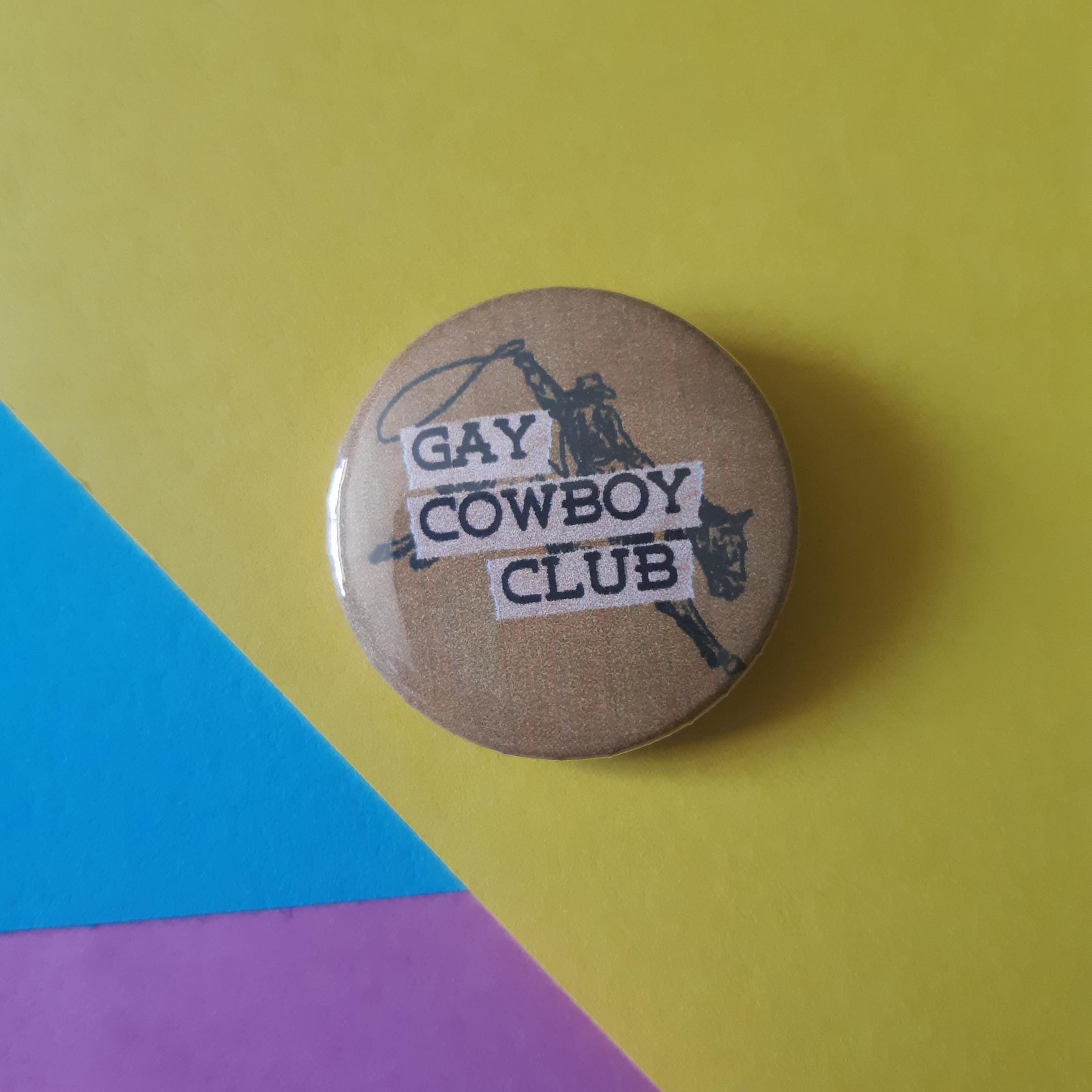 A round pinback button badge with a brown kraft-paper background and black western-themed graphic, featuring the text 'GAY COWBOY CLUB' on white strips. The badge is photographed on a colourful background split diagonally into yellow, blue, and pink sections.