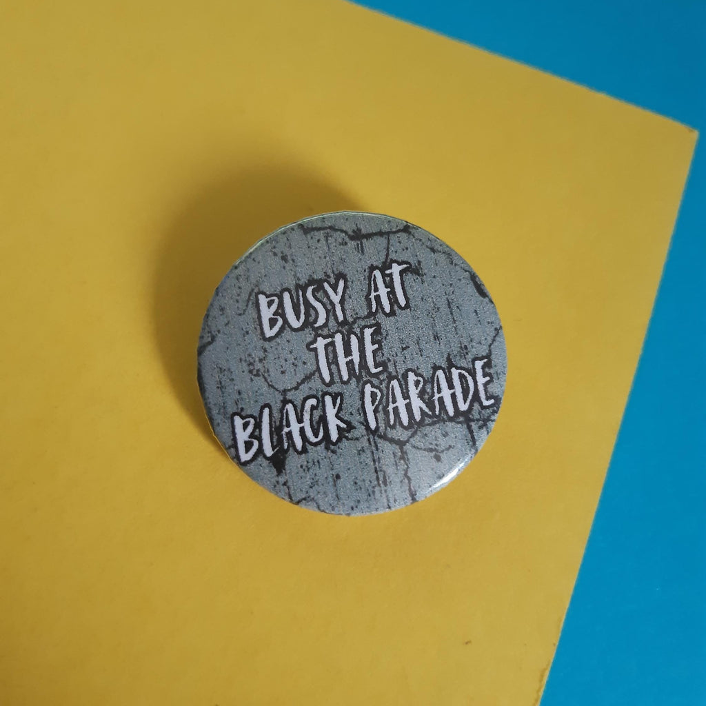 A round pin badge with a distressed gray background and white text reading 'BUSY AT THE BLACK PARADE', placed on a yellow sheet of paper with a blue background visible in the upper right corner.
