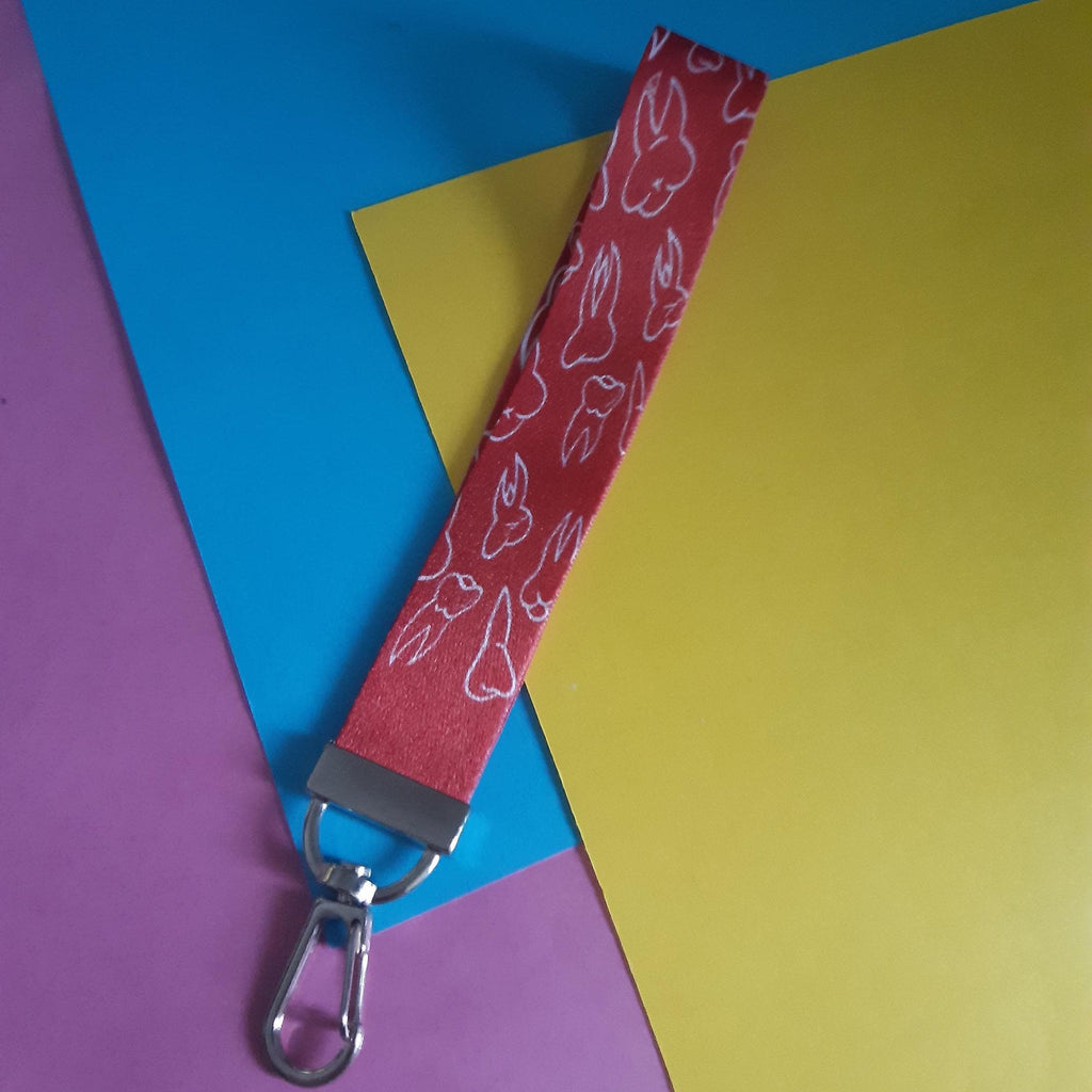 A fabric wristlet strap with a red background and a white teeth pattern, attached to a silver metal swivel clasp. The strap is laid diagonally across a background composed of three colored paper sheets: pink, blue, and yellow.