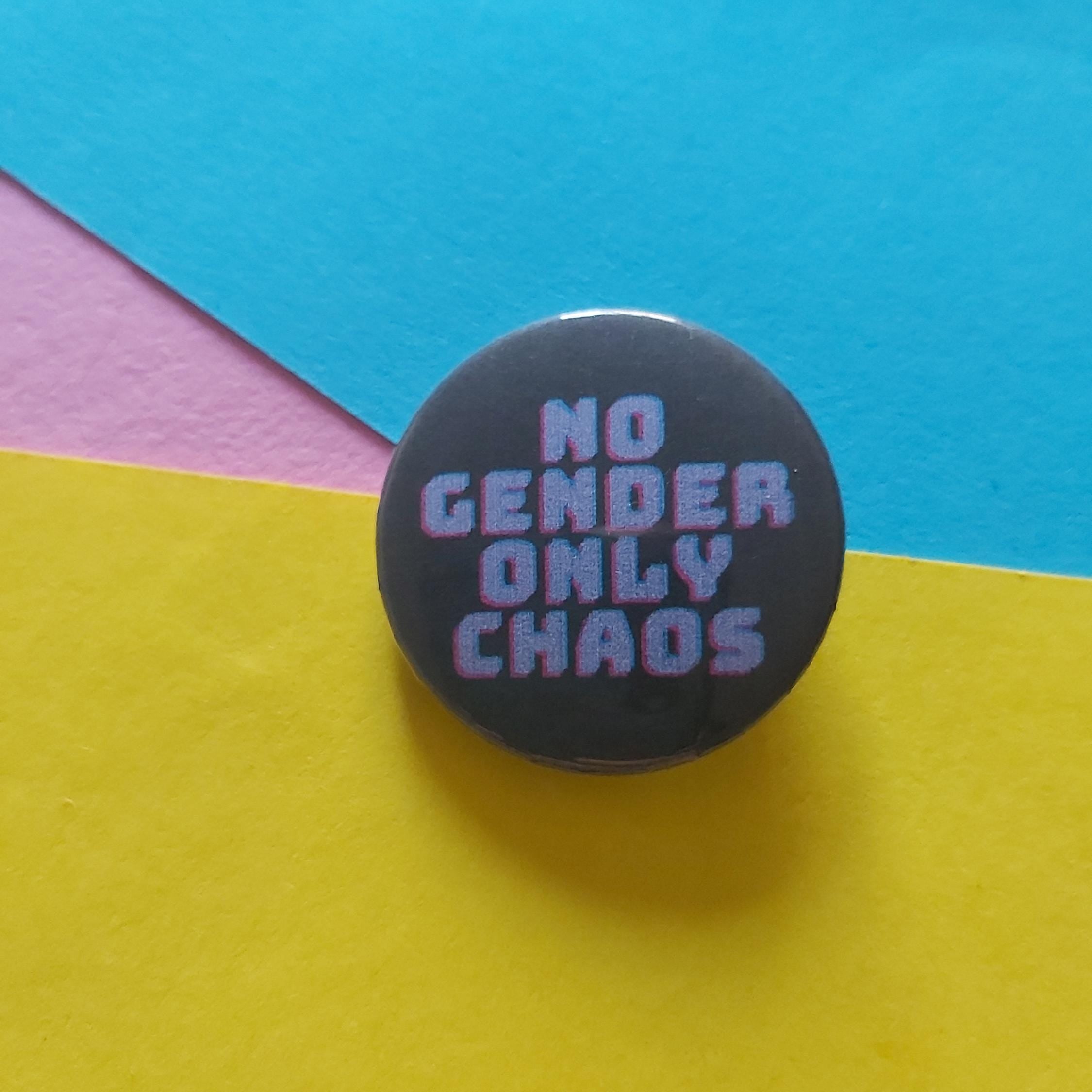 A round pinback button with the text 'NO GENDER ONLY CHAOS' in purple bubble letters on a black background, placed on a colourful paper backdrop of yellow, blue, and pink.