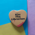 A heart-shaped pin with the text 'Kiss The Librarian' in black, featuring red blood-splatter details on a yellow background, set against a colourful background of blue, purple, and yellow paper.
