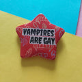A red star-shaped pin with the text 'VAMPIRES ARE GAY' in black letters on a white banner, decorated with white line drawings of vampire fangs, set against a yellow and blue background.