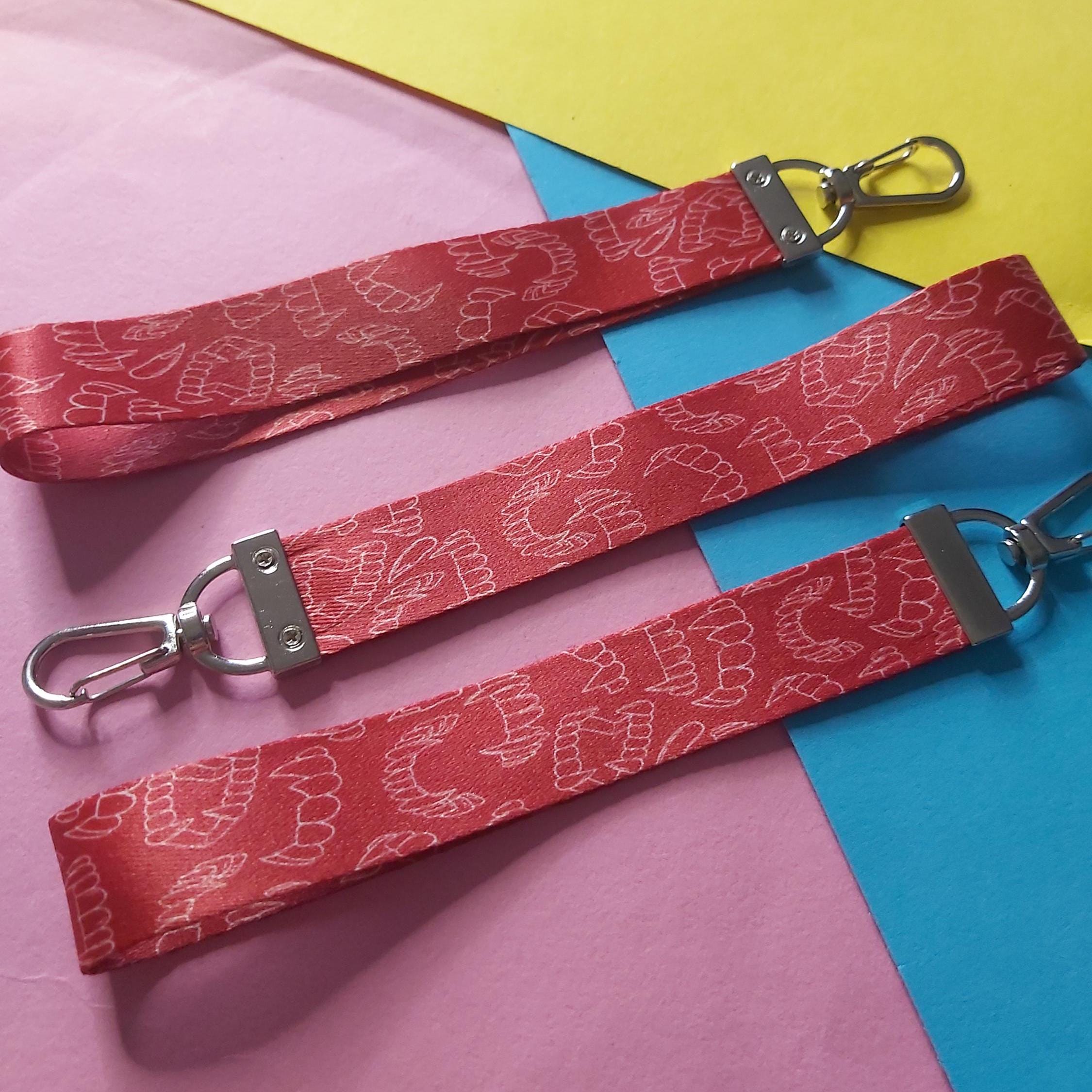 Three red fabric lanyards with a white line-art pattern of vampire fangs and silver metal swivel clips, laid diagonally on a colourful background of pink, yellow, and blue paper.