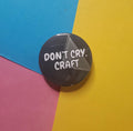 A round pin badge with the text 'DON'T CRY, CRAFT' in white lettering on a dark gray background, placed on a diagonally split blue, pink, and yellow paper backdrop.