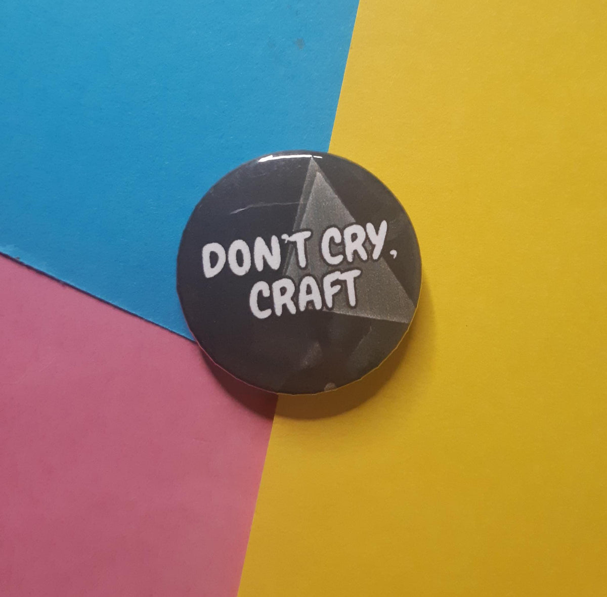 A round pin badge with the text 'DON'T CRY, CRAFT' in white lettering on a dark gray background, placed on a diagonally split blue, pink, and yellow paper backdrop.