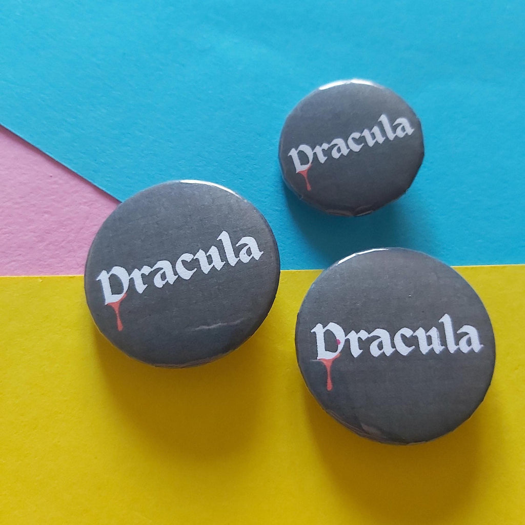Three round black pin-back buttons with the word 'Dracula' in white gothic lettering and a small red blood-drip accent, arranged on a colorful background of cyan, pink, and yellow paper.