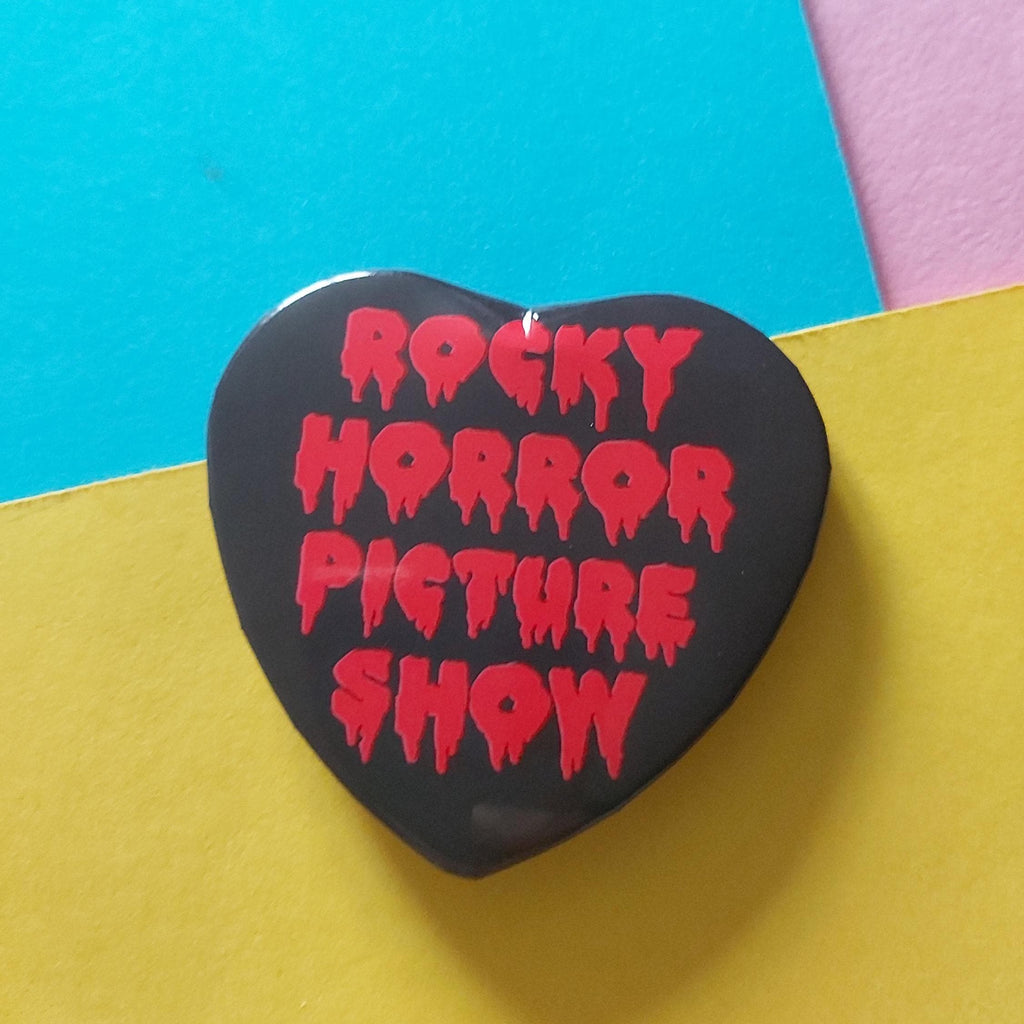 A heart-shaped black pin badge with the words 'ROCKY HORROR PICTURE SHOW' in dripping red lettering, photographed on a colourful background of cyan, yellow, and pink paper.
