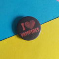 A round pin-back button badge with the text 'I [heart] VAMPIRES' in red, placed on a diagonally split blue and yellow background.