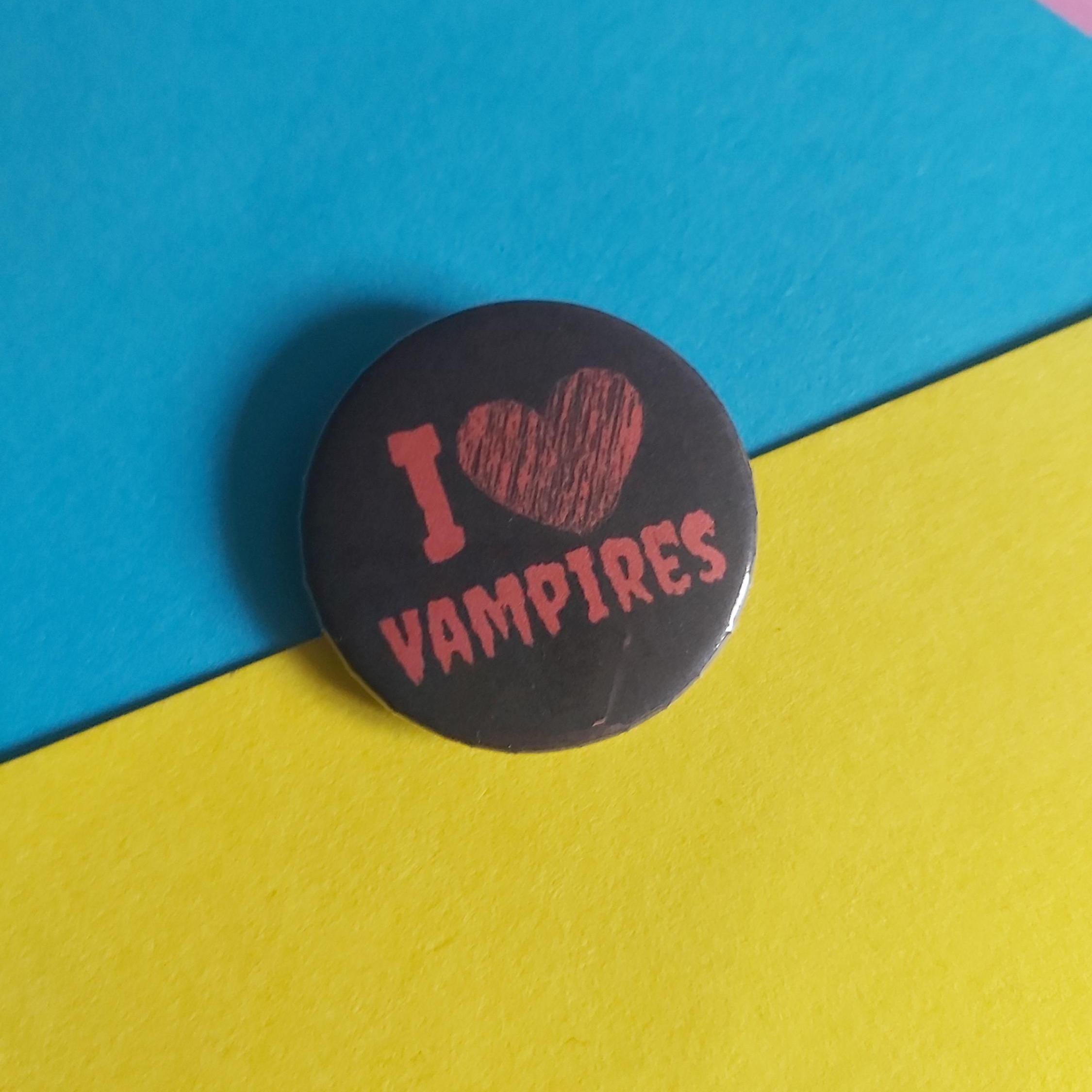 A round pin-back button badge with the text 'I [heart] VAMPIRES' in red, placed on a diagonally split blue and yellow background.
