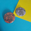 Two round pin-back buttons with the text 'QUEER CODED VILLAIN' on a bright blue and yellow background.