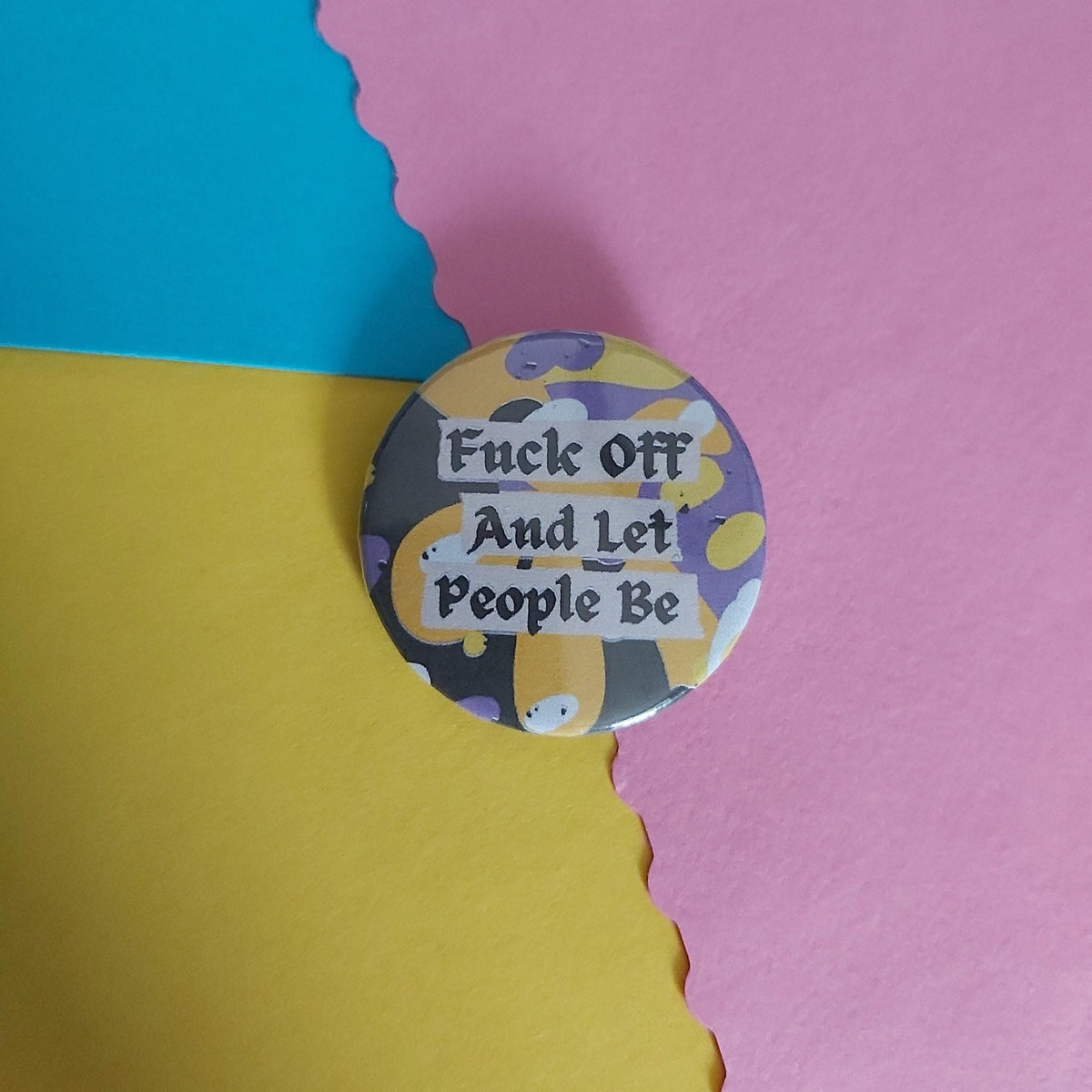 A round pinback button badge with a colourful abstract background and the text 'Fuck Off And Let People Be' in black gothic lettering, displayed on a background split diagonally into blue, yellow, and pink sections.