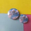 Two round pin-back buttons with a sky-and-cloud print and the text 'Fuck ai art' in dark lettering, displayed on a colorful pastel background.