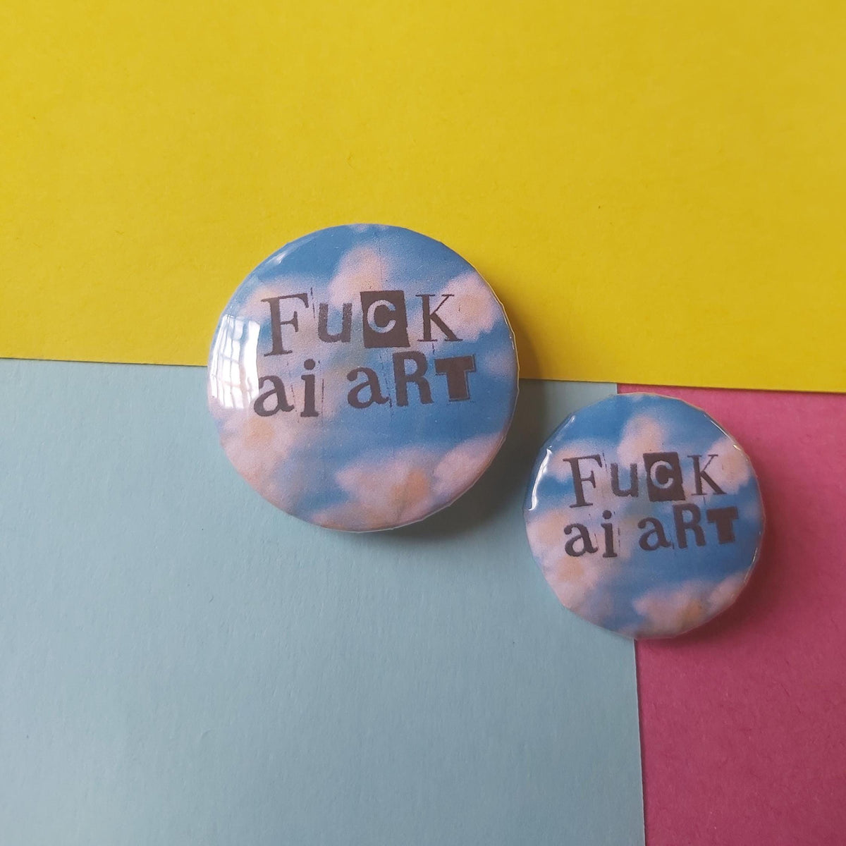 Two round pin-back buttons with a sky-and-cloud print and the text 'Fuck ai art' in dark lettering, displayed on a colorful pastel background.