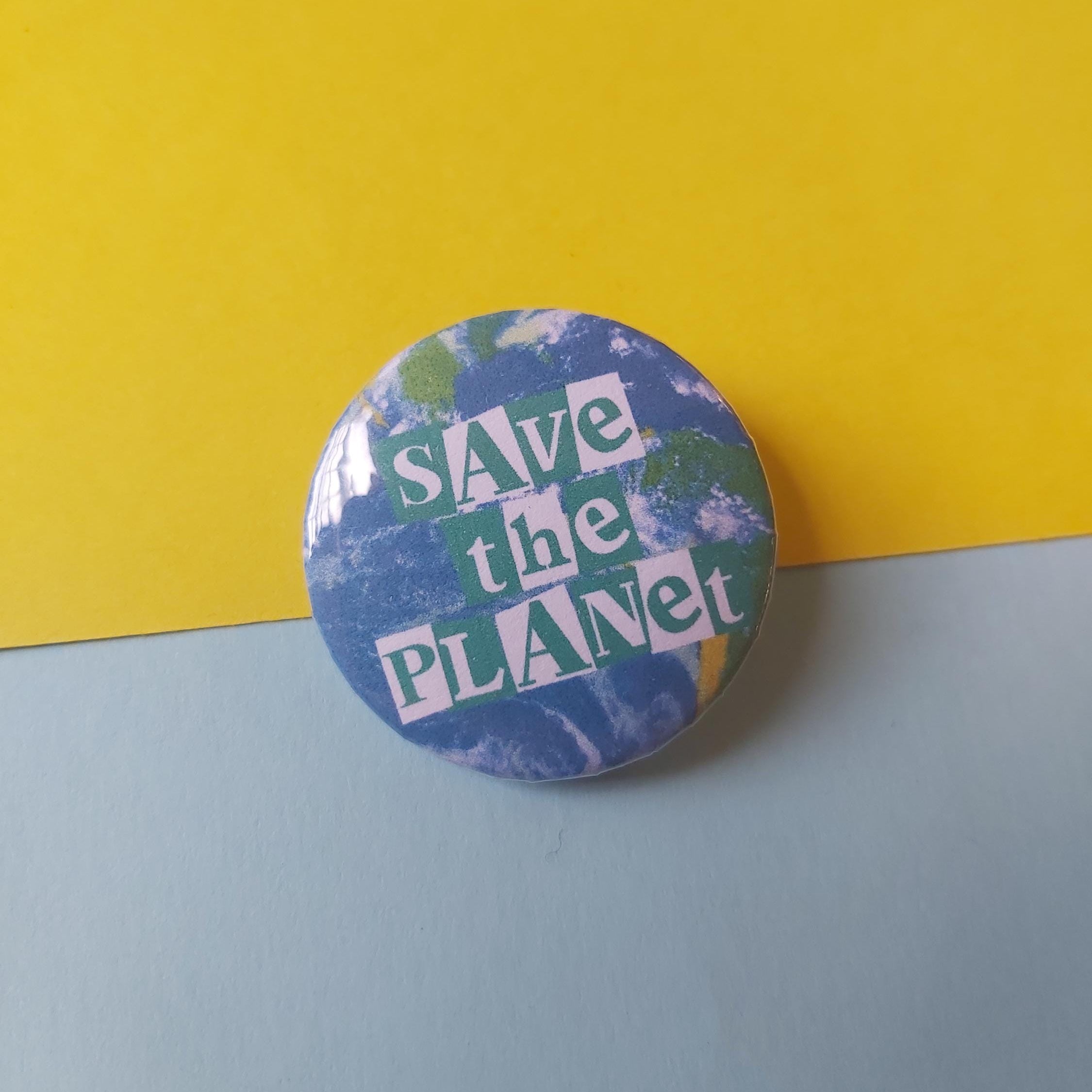 A round pin badge featuring a blue and green Earth design with the text 'SAVE the PLANET' in white and green, placed on a split yellow and light blue background.