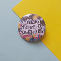 A round pinback button badge featuring the text 'PUNKS RESPECT PRONOUNS' in a black block font over a colourful abstract paint splatter background, placed on a light blue surface with a yellow paper corner behind it.