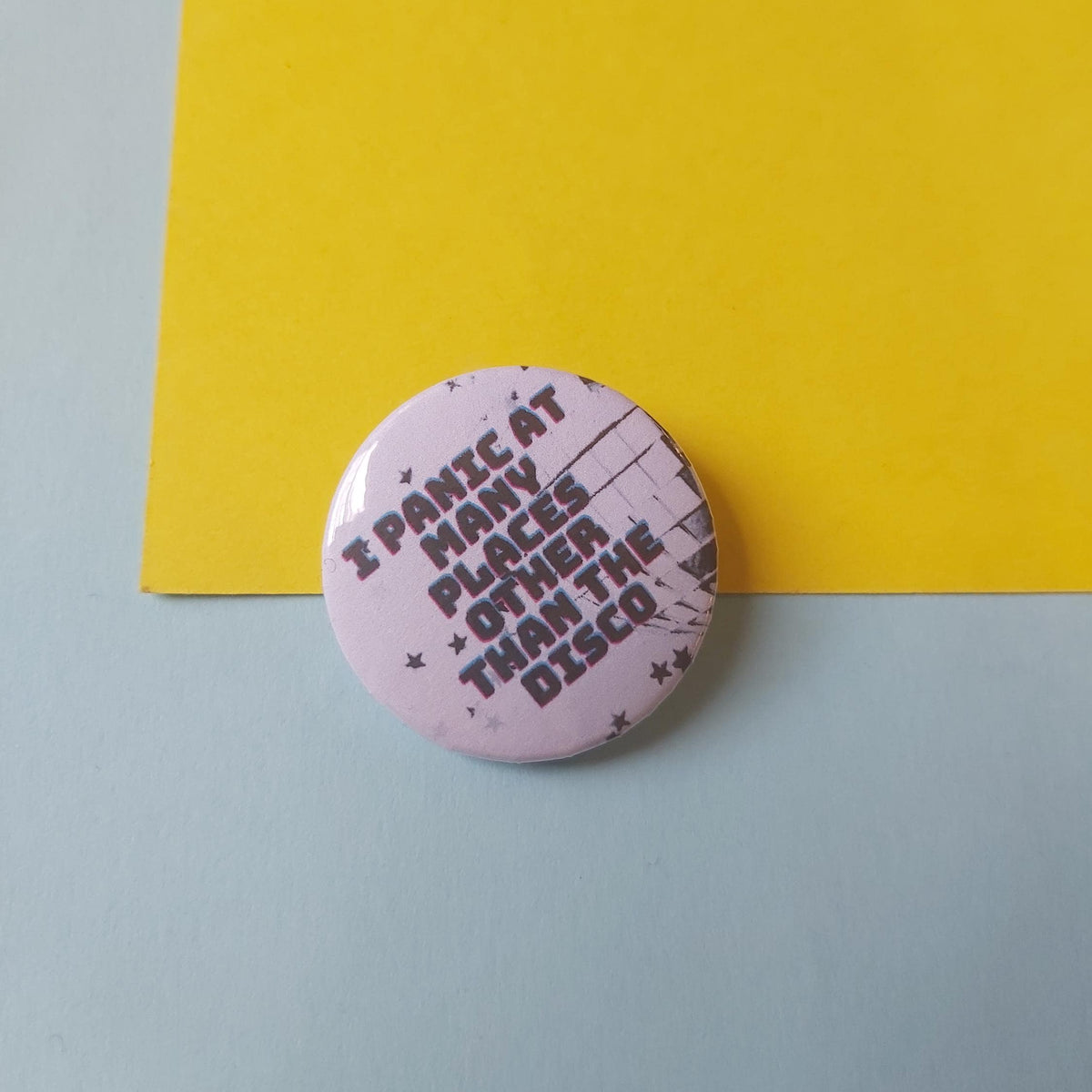 A round pin-back button badge featuring the text 'I PANIC AT MANY PLACES OTHER THAN THE DISCO' in dark lettering on a light pink background, placed on a light blue surface with a bright yellow paper sheet behind it.