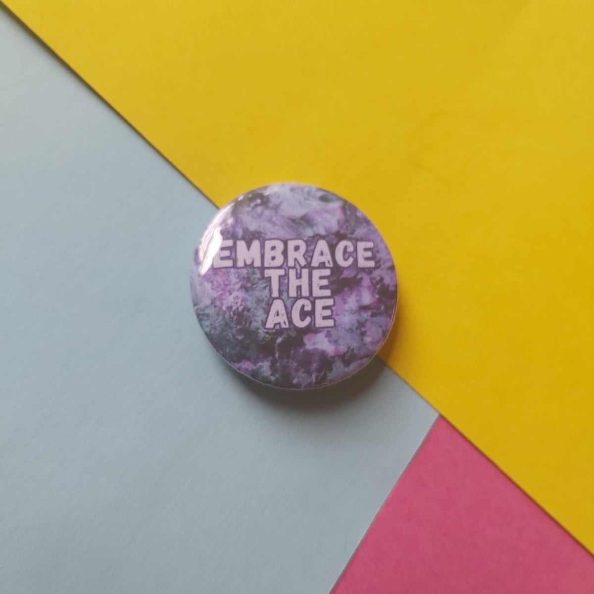 A round pin badge with a purple and grey marble pattern and the text 'EMBRACE THE ACE' in white, resting on a diagonally split yellow, light grey, and pink background.