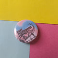 A round pinback button badge featuring a black dinosaur illustration and the text 'MAKE TRANSPHOBIA EXTINCT' on a pink and blue gradient background, placed on a three-colored paper surface with yellow, light blue, and pink sections.