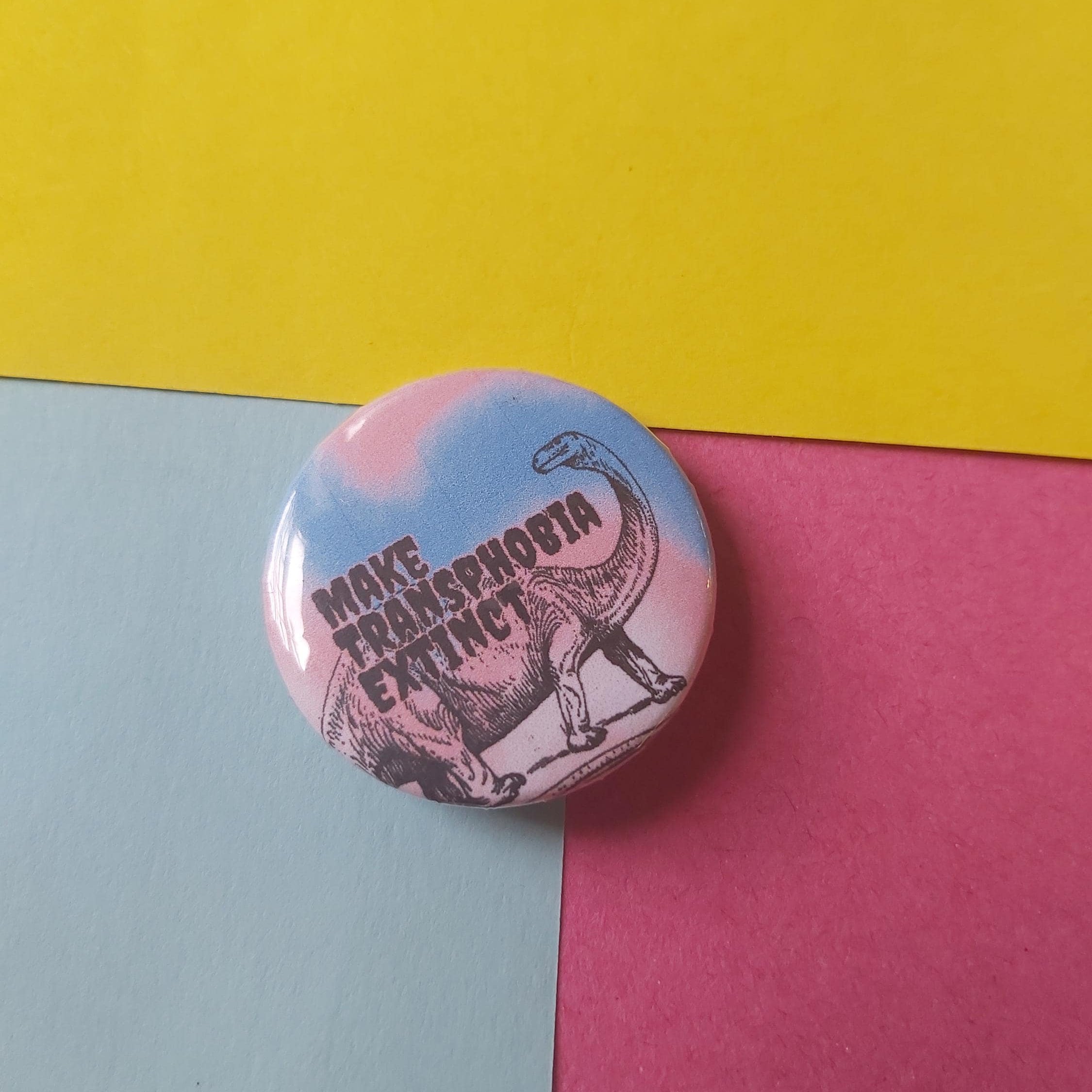 A round pinback button badge featuring a black dinosaur illustration and the text 'MAKE TRANSPHOBIA EXTINCT' on a pink and blue gradient background, placed on a three-colored paper surface with yellow, light blue, and pink sections.