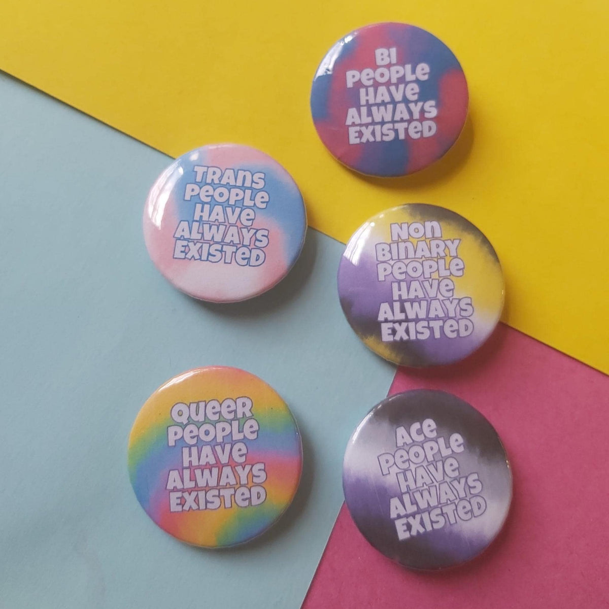 Five round pinback buttons with pride-themed slogans reading 'BI PEOPLE HAVE ALWAYS EXISTED', 'TRANS PEOPLE HAVE ALWAYS EXISTED', 'NON BINARY PEOPLE HAVE ALWAYS EXISTED', 'QUEER PEOPLE HAVE ALWAYS EXISTED', and 'ACE PEOPLE HAVE ALWAYS EXISTED'. Each button has a different pastel rainbow tie-dye background and white text.