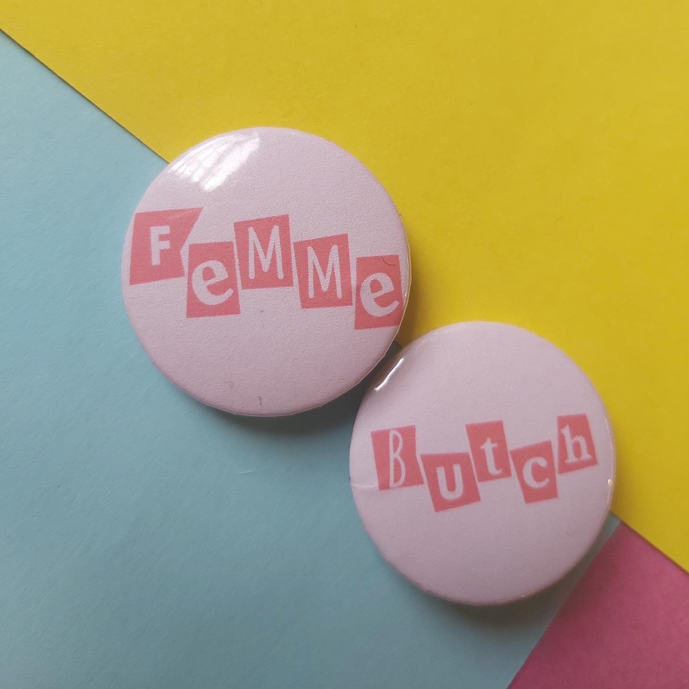 Two round pinback buttons with pink lettering on a pastel background. One button reads 'Femme' and the other reads 'Butch' in pink blocks.
