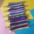 Colourful striped lanyards with silver metal lobster clasps, arranged diagonally on a background of pink, yellow, and blue paper.