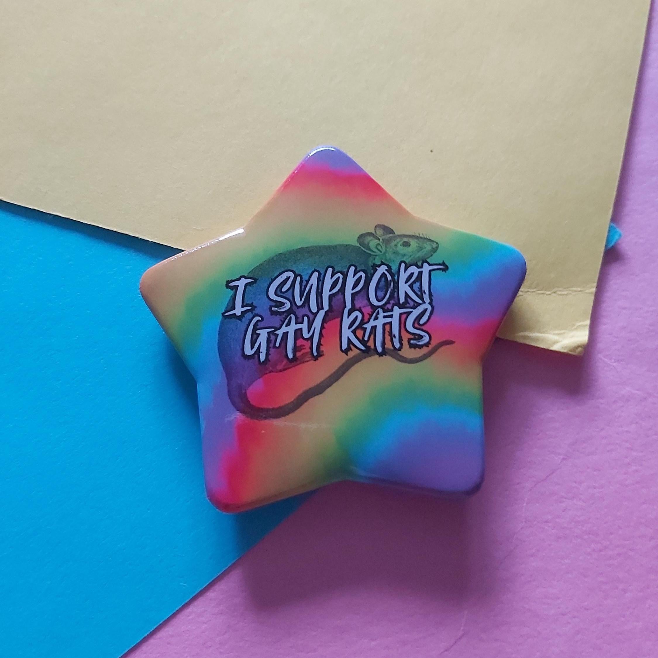 A star-shaped pin badge featuring a rainbow tie-dye pattern and the text 'I SUPPORT GAY RATS', displayed on a split blue and purple background with a yellow paper sheet.