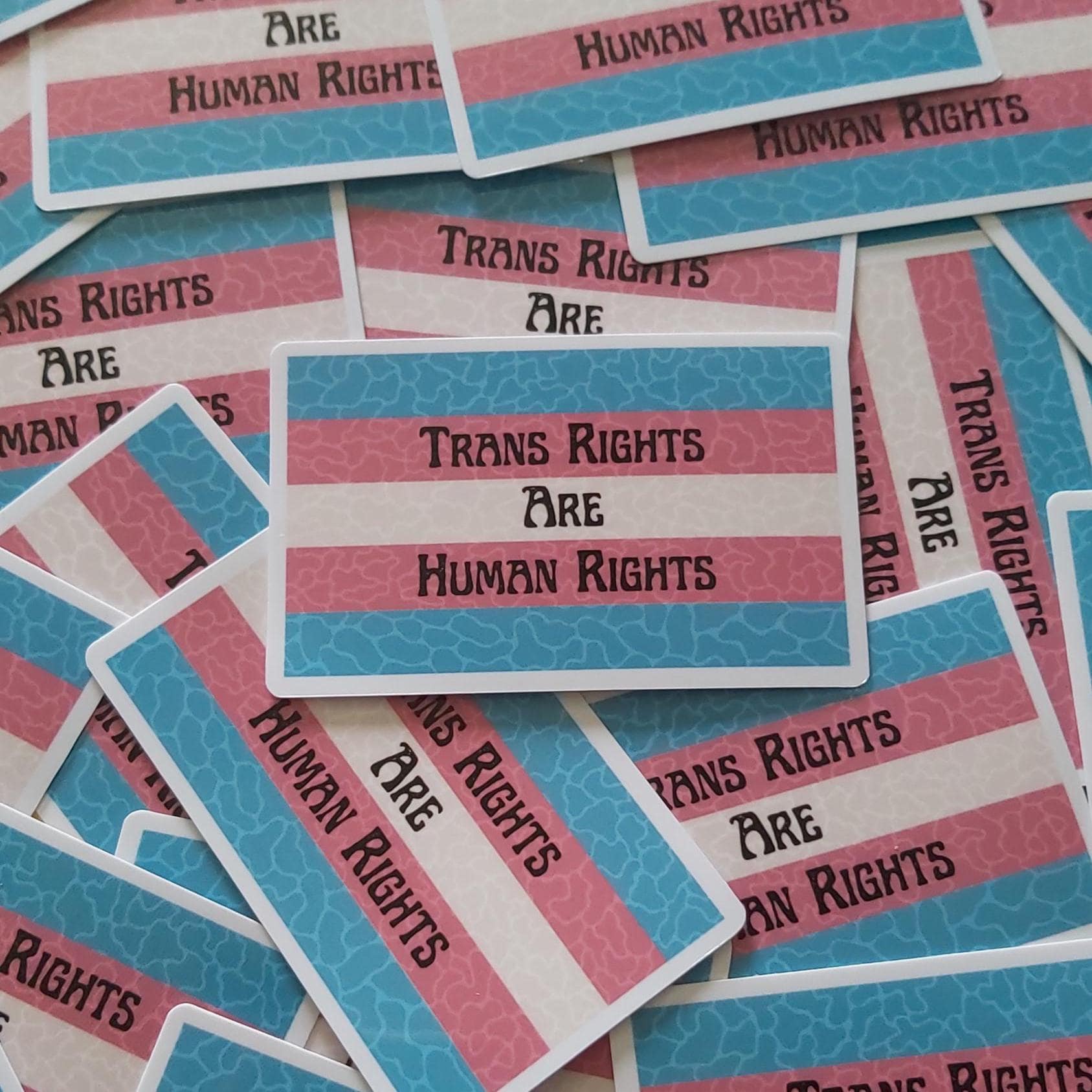 A pile of rectangular stickers with the phrase 'Trans Rights Are Human Rights' printed in black text over a pink, white, and blue striped transgender flag.