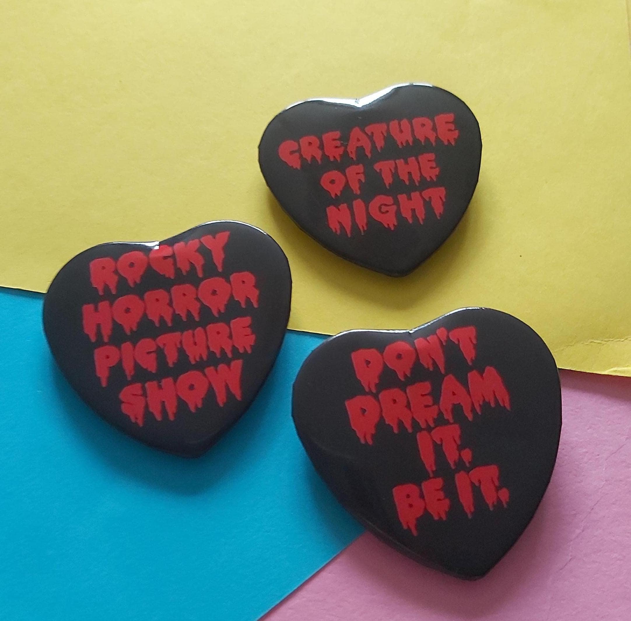Three heart-shaped black pins with red horror-themed text, displayed on a colourful background of yellow, blue, and pink paper.