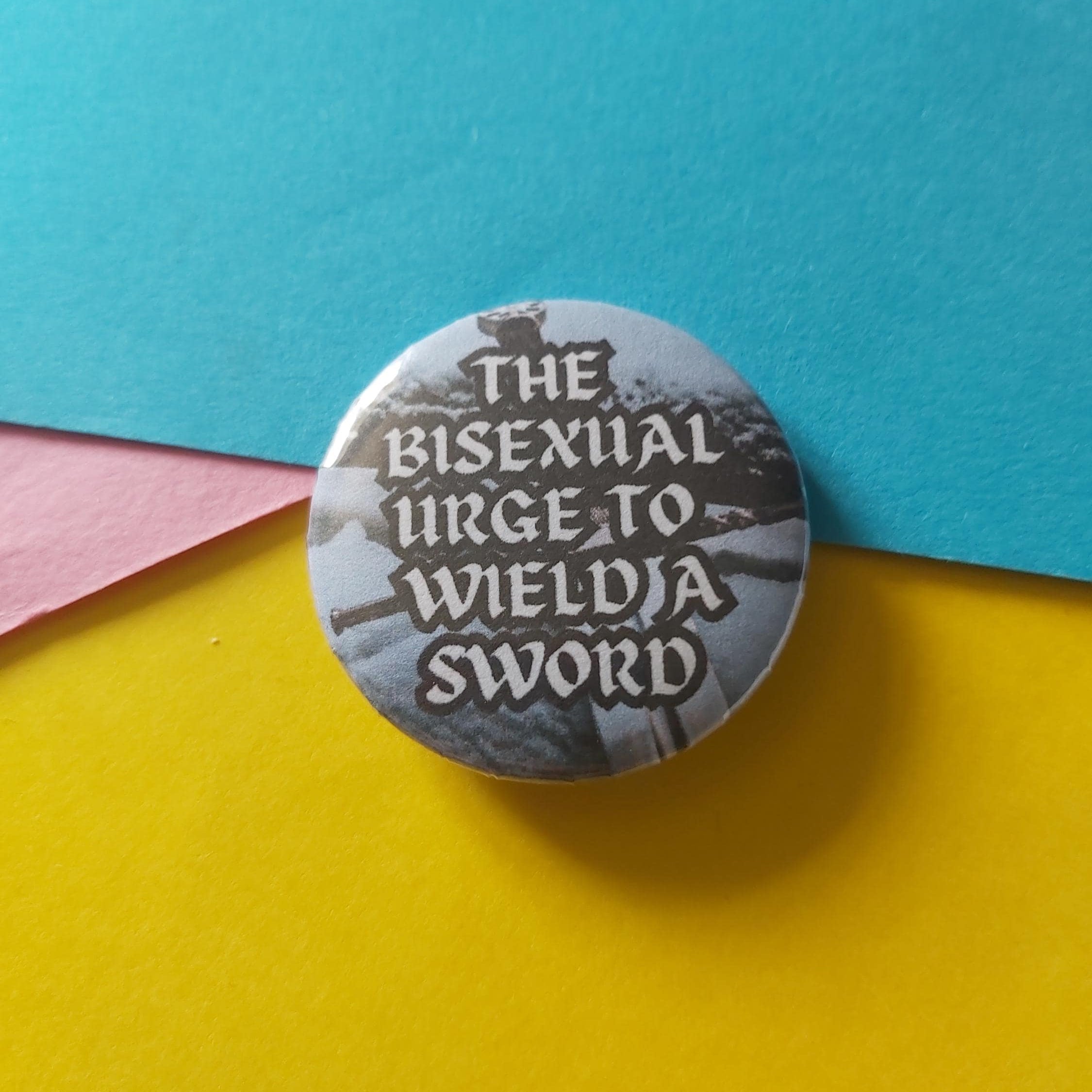 A round pinback button featuring the text 'THE BISEXUAL URGE, TO WIELD A SWORD' in white lettering over a dark fantasy scene
