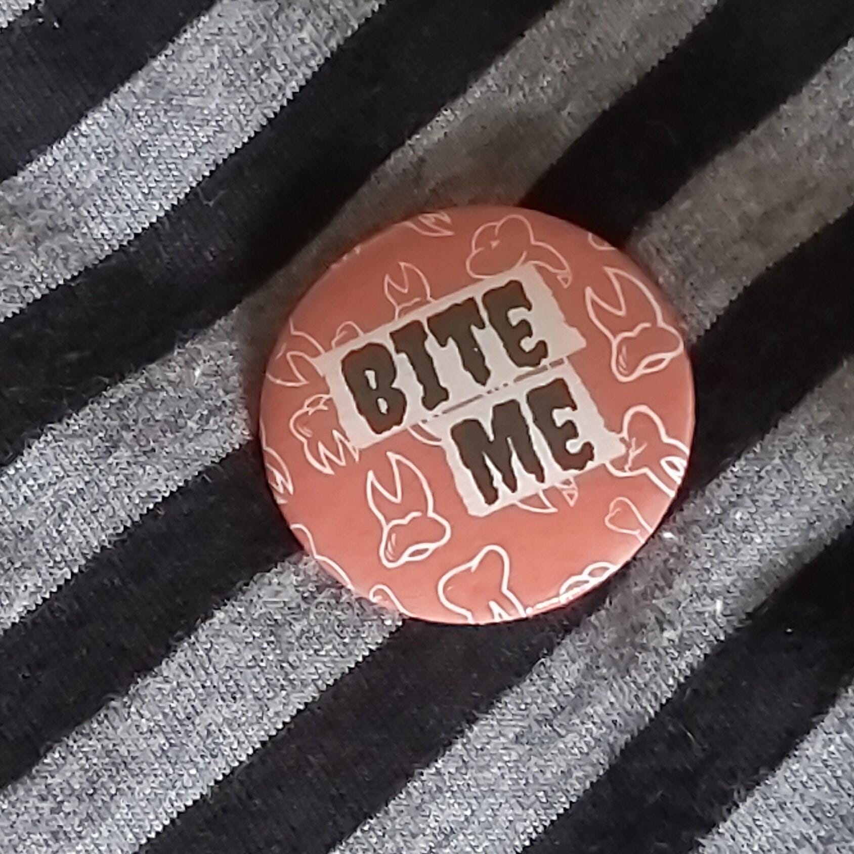 A round pin badge with a red background, featuring the words 'BITE ME' in black letters on white cutout shapes, surrounded by white teeth line drawings.