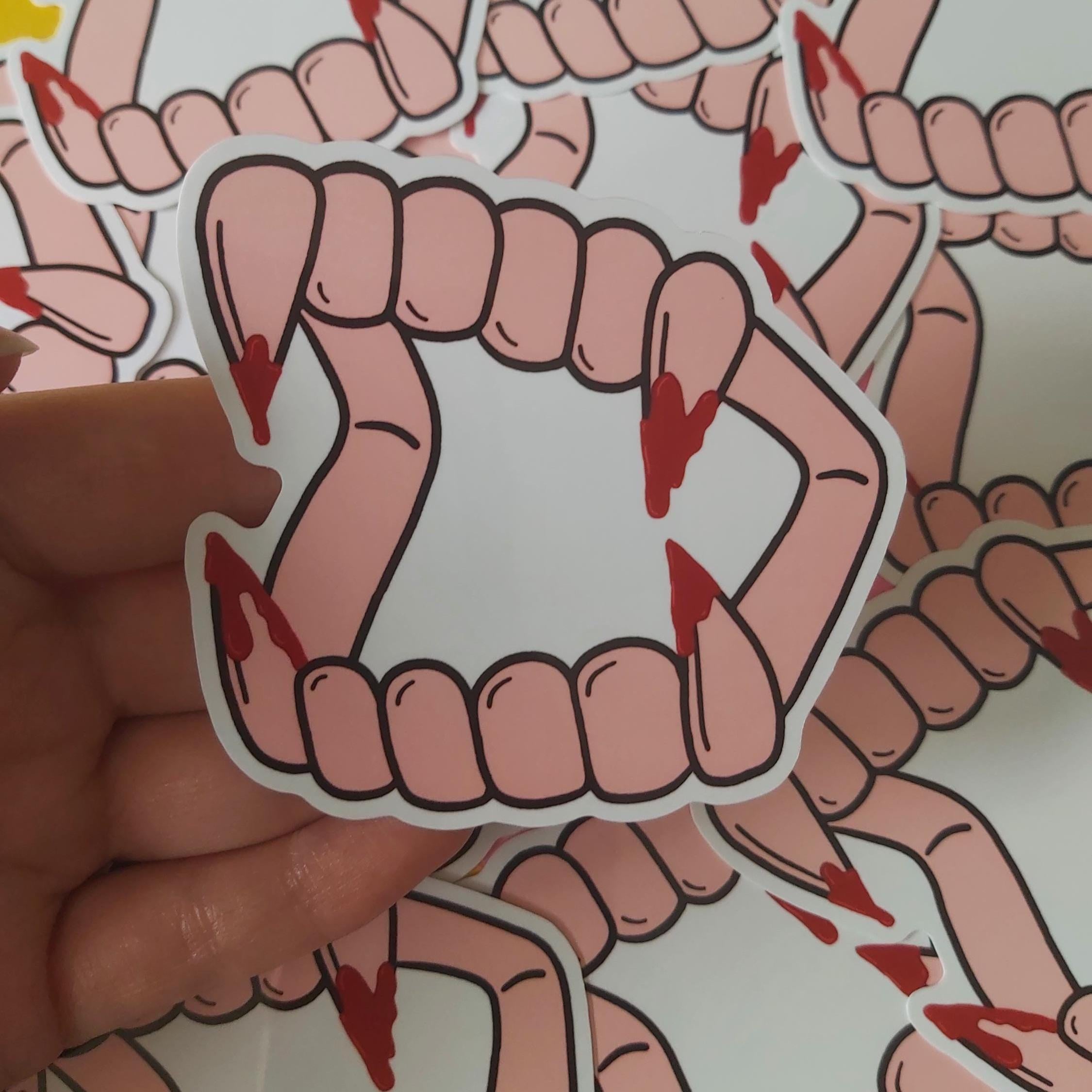 A hand holds a die-cut sticker depicting vampire fangs with black outlines and red blood details, with other identical stickers scattered in the background.