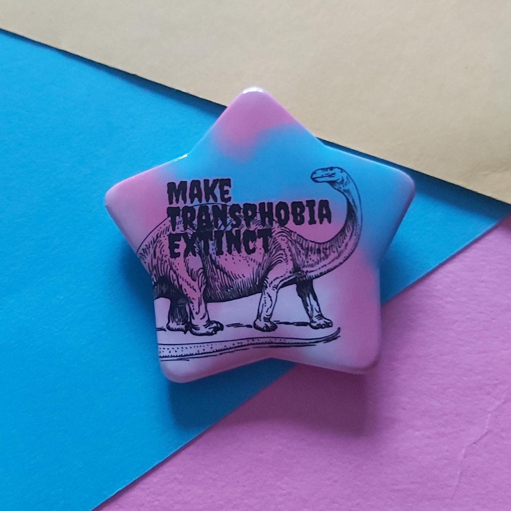 A star-shaped pin featuring a black line art illustration of a long-necked dinosaur and the text 'MAKE TRANSPHOBIA EXTINCT' in black lettering. The pin has a pink and light blue tie-dye gradient background.