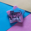 A star-shaped pin featuring a black line art illustration of a long-necked dinosaur and the text 'MAKE TRANSPHOBIA EXTINCT' in black lettering. The pin has a pink and light blue tie-dye gradient background.