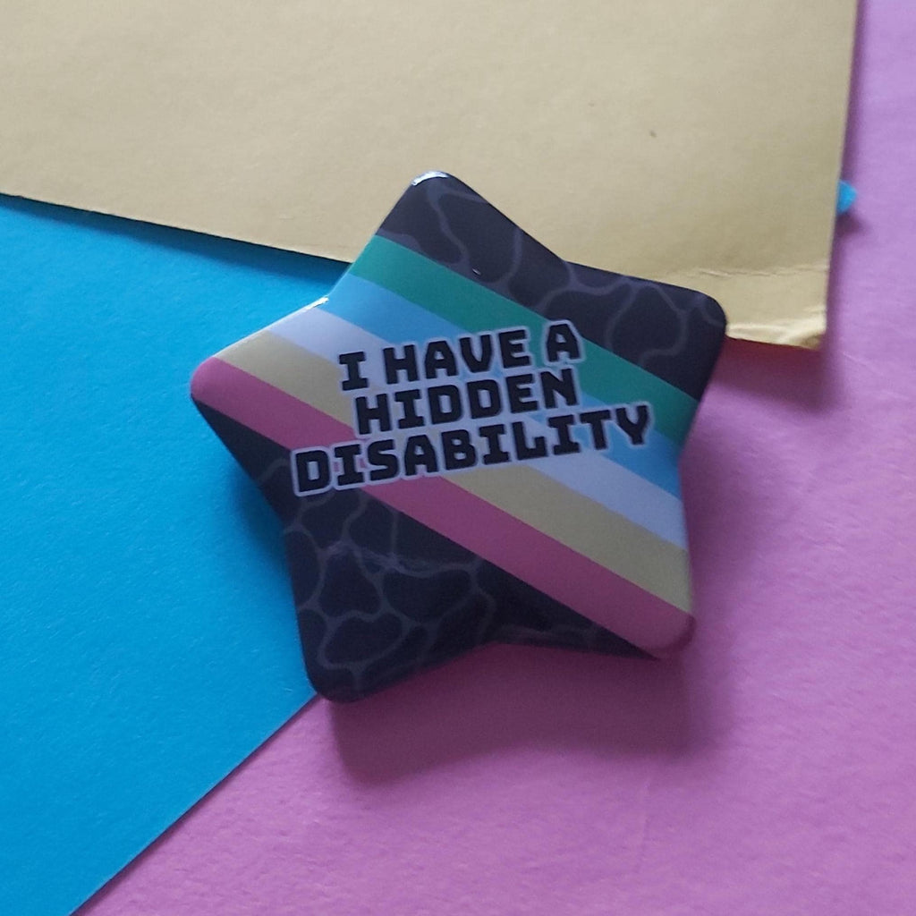A star-shaped pin badge with the text 'I HAVE A HIDDEN DISABILITY' in white, outlined lettering. The badge features a dark patterned background with diagonal stripes in teal, light blue, pink, and beige.