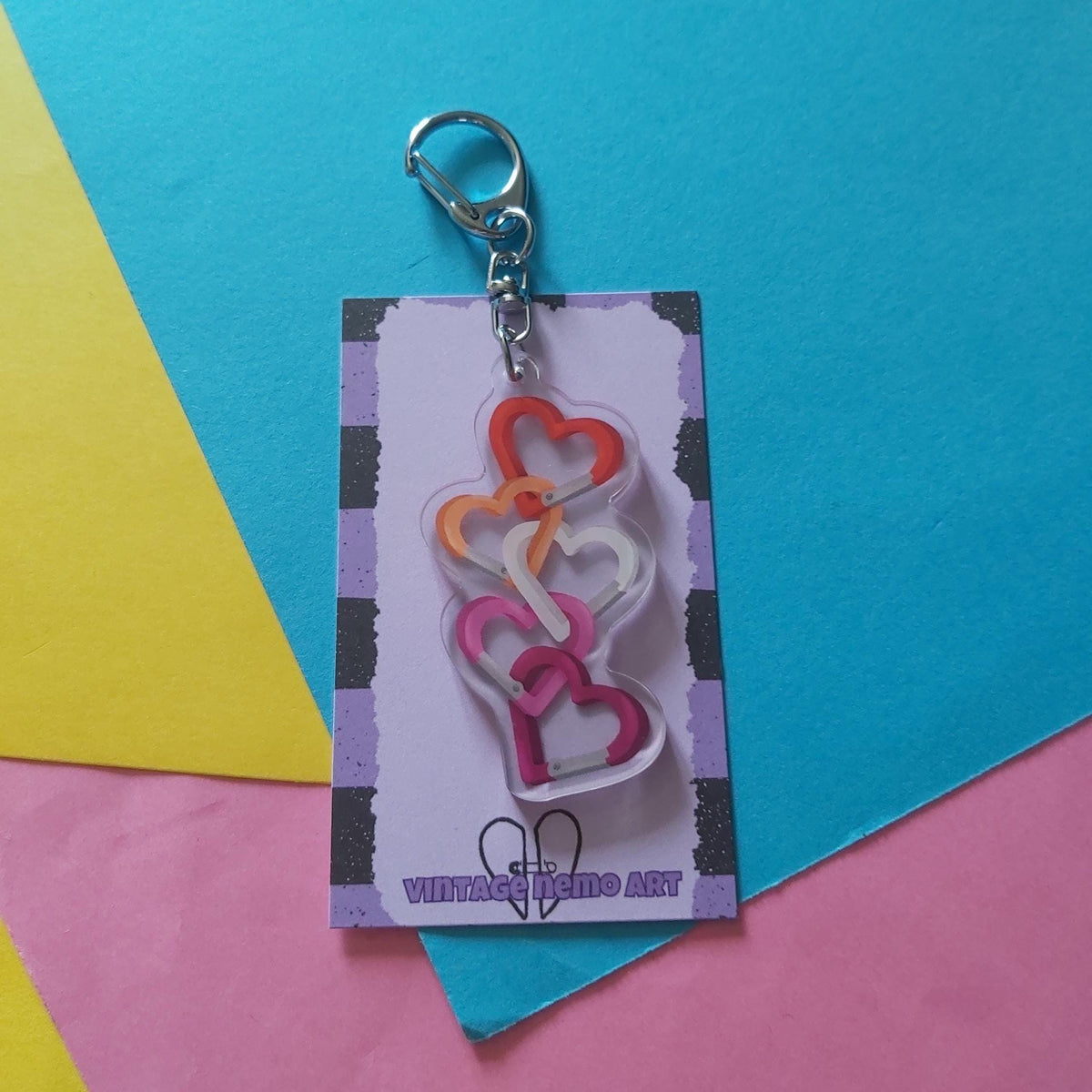 A keychain featuring five interlocking heart carabiners in orange, yellow, white, and pink, attached to a silver clasp and displayed on a branded backing card.