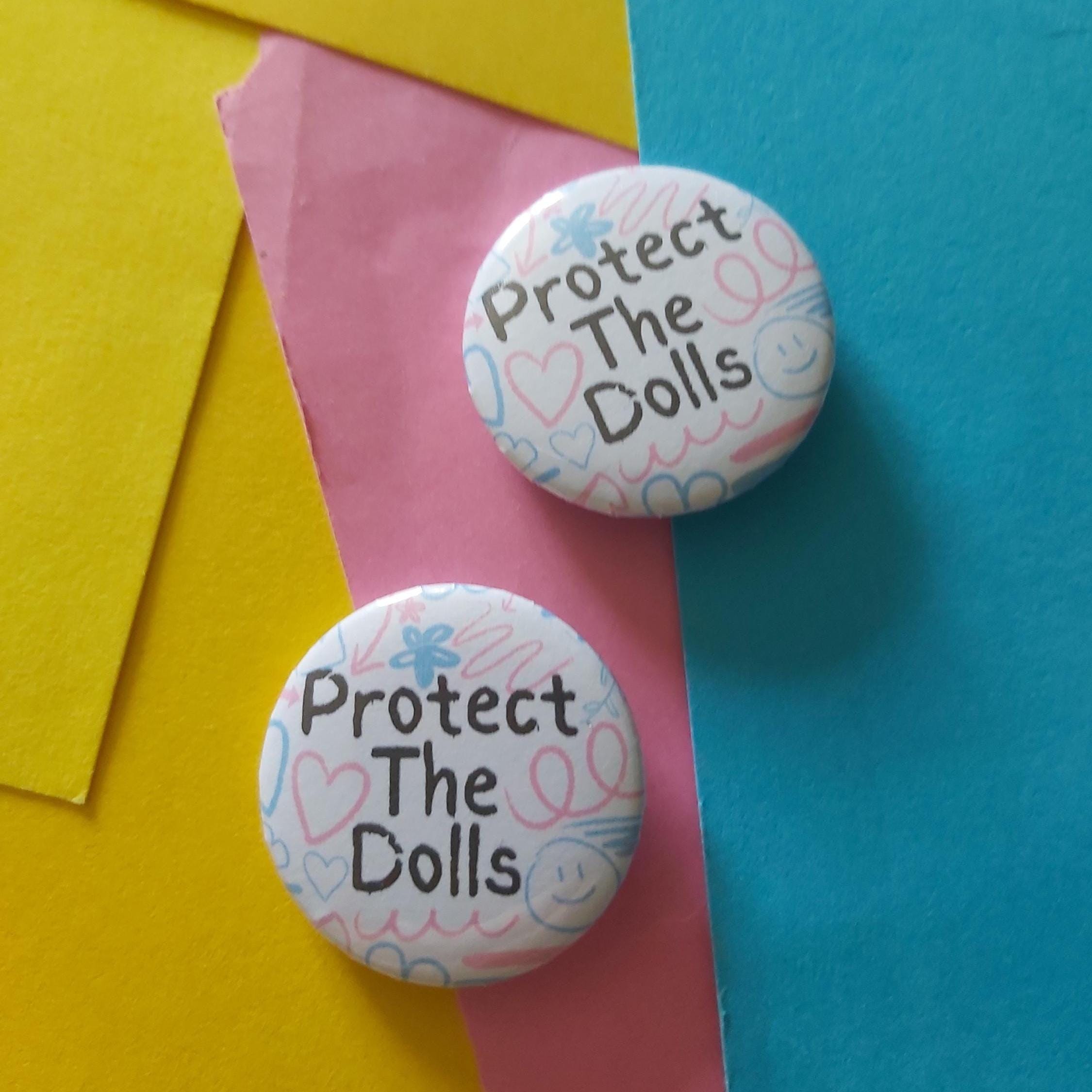 Two round pin-back buttons with the text 'Protect The Dolls' and a pastel doodle pattern, displayed on a colorful background of yellow, pink, and blue paper.