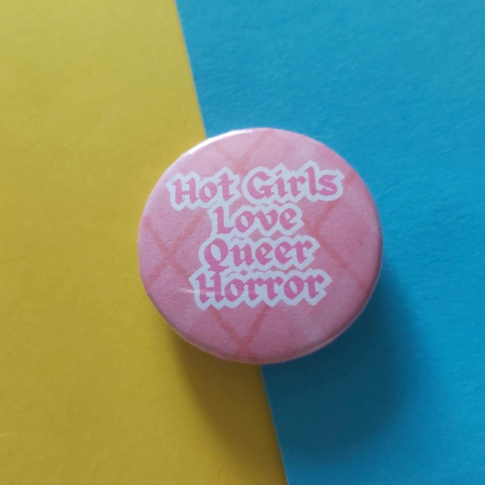 A round pink pinback button with the text 'Hot Girls Love Queer Horror' in white gothic lettering, set against a diagonally split yellow and blue background.