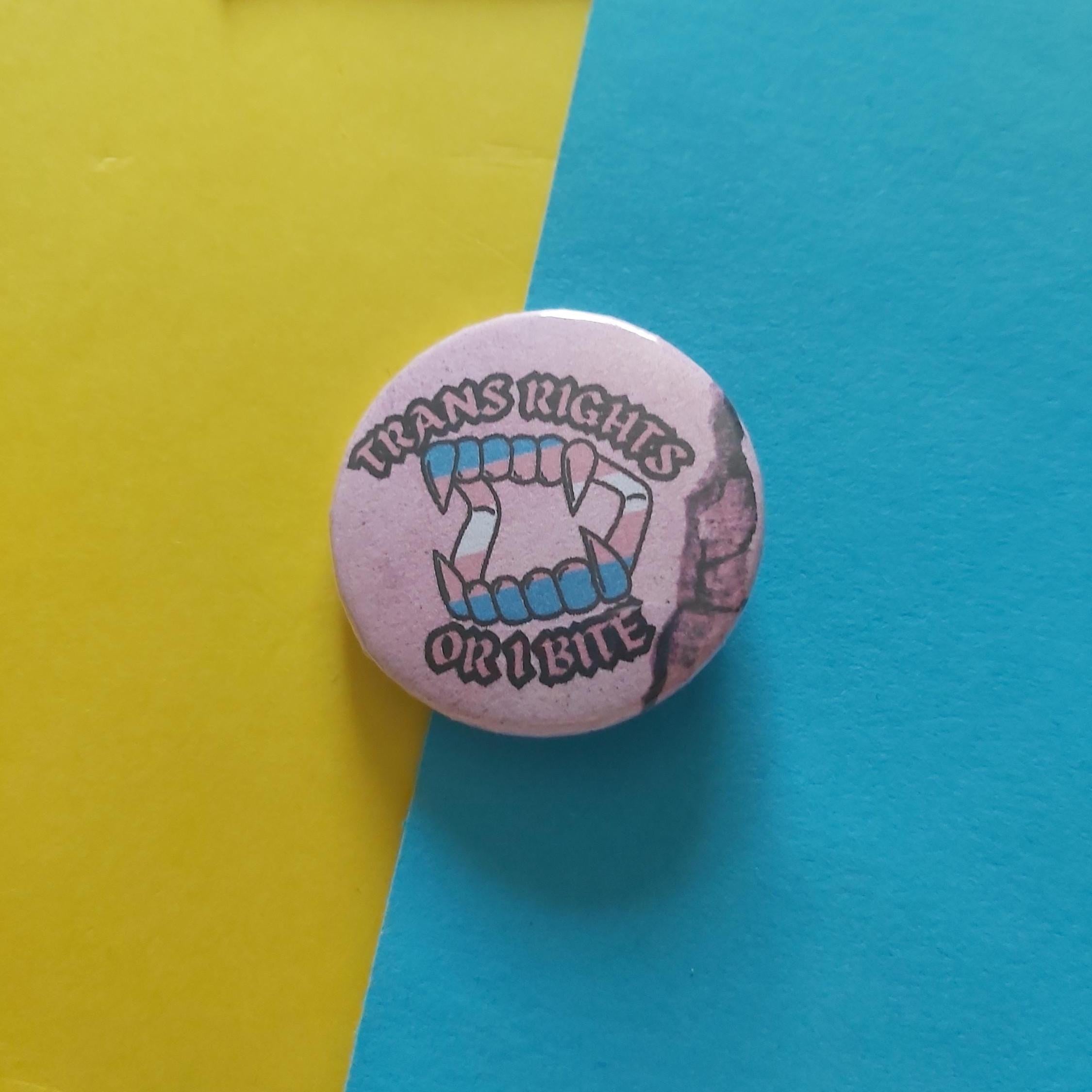 A round pinback button badge featuring a graphic of vampire teeth with blue accents and the text 'TRANS RIGHTS OR BITCHES' in black gothic lettering, set against a diagonally split yellow and blue background.